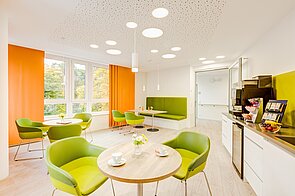Blick in die große, helle Aufenthaltslounge mit Tischen und Stühlen in grün und weiß, orangenen Vorhängen und Kaffeetheke